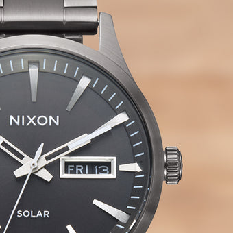Close-up of a dark grey Nixon solar watch with a black face, metallic hands, and a date display showing "FRI 13".