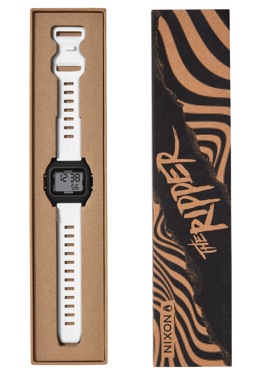 A white Nixon digital watch with a black face and white strap is presented in an open brown cardboard box next to its black and brown striped packaging that reads "THE RIPPER" and "NIXON". View 10